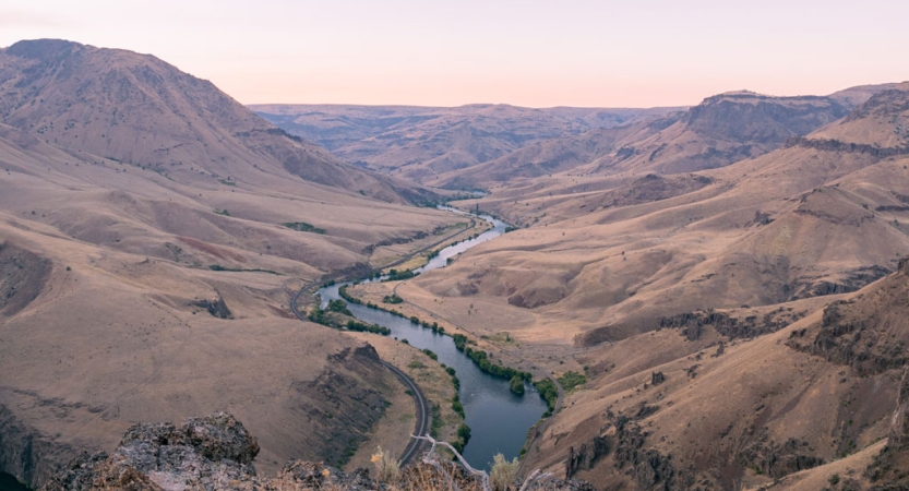 From an aerial point of view, a river moves through a desert mountainous landscape under pink and purple skies. 